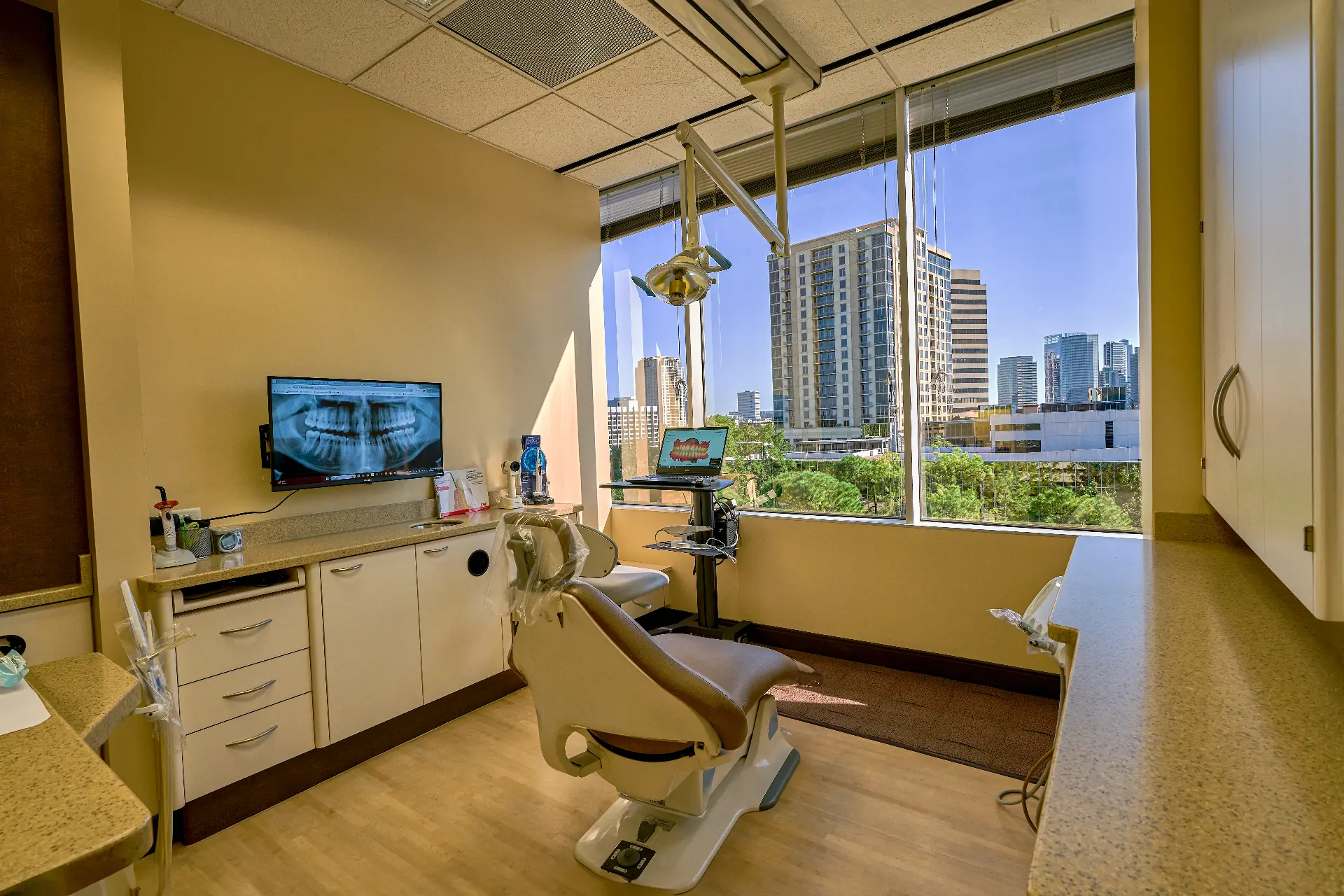 Sunlit dental treatment room at Uptown Dentistry with a panoramic X-ray on screen and floor-to-ceiling views of Uptown Houston